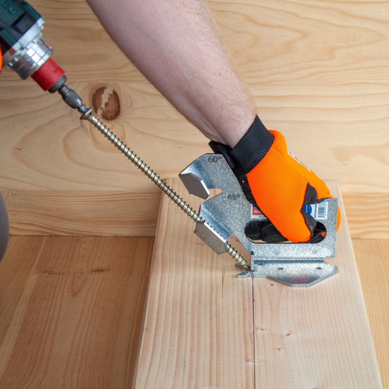 Die GSCREW-Schraubenführung in Aktion: Von links kommend, dreht ein automatischer Schrauber eine sehr lange Schraube im schrägen Winkel in ein Stück Holz ein. Die Schraube wird durch eine GSCREW geführt, festgehalten von einer Hand in einem orangenen Handschuh.